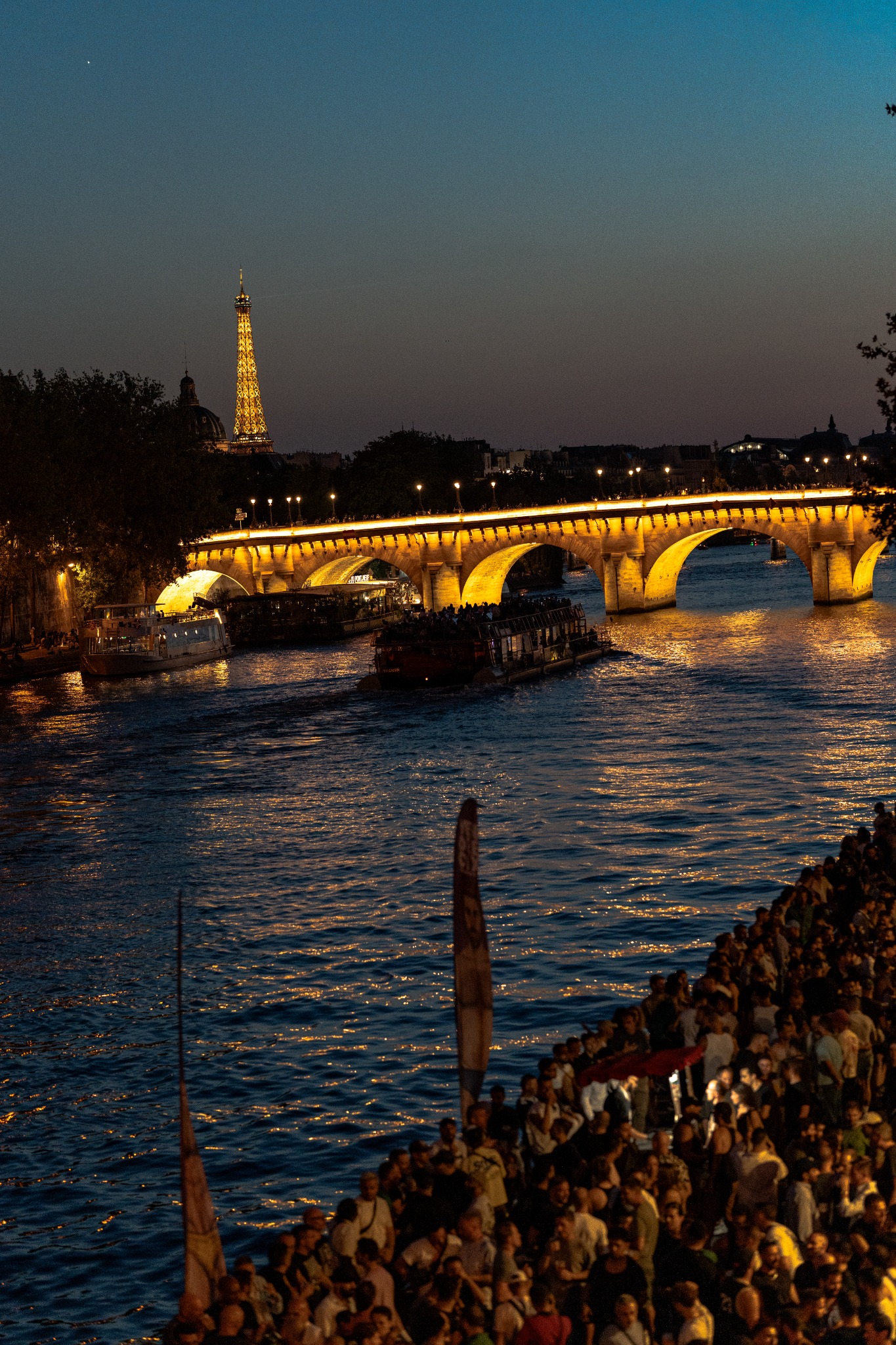 Crowd gathered beside the Seine beneath the illuminated Eiffel Tower